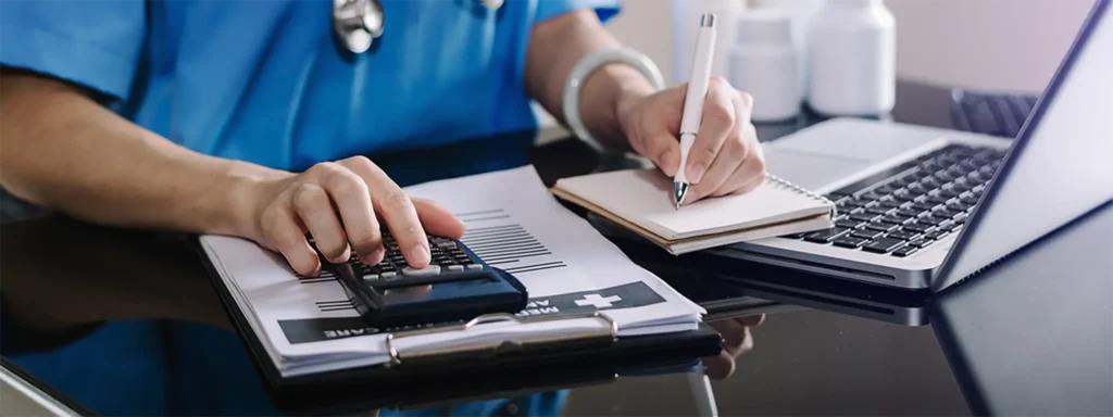 A doctor writes notes on paper while using a calculator on the table beside them.