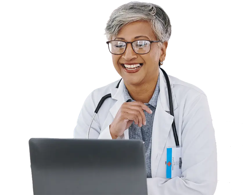 A smiling woman in a white lab coat sits at a laptop, engaged in her work.